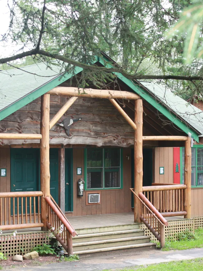 A log-trimmed cabin with a green roof, front porch and steps, shaded by trees