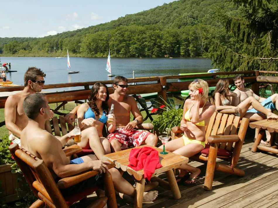 Adults in swimsuits relaxing around a table on a wooden deck overlooking the lake and sailboats
